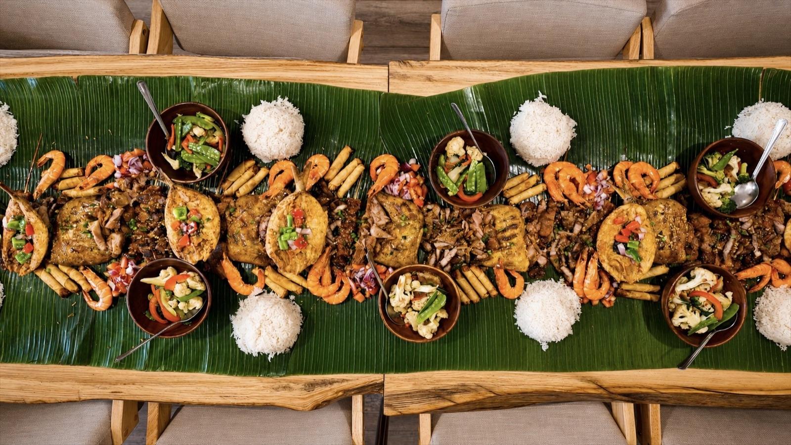 An overhead IPK Kamayan boodle fight spread laid end to end on banana leaves — inasal, crispy pata, shrimp, lumpia and mounds of rice.